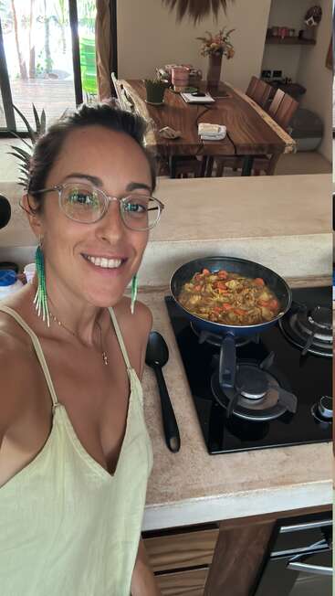 Una mujer con gafas y pendientes verdes sonríe mientras prepara un plato de verduras en una sartén sobre la cocina. Al fondo se ve un acogedor comedor.
