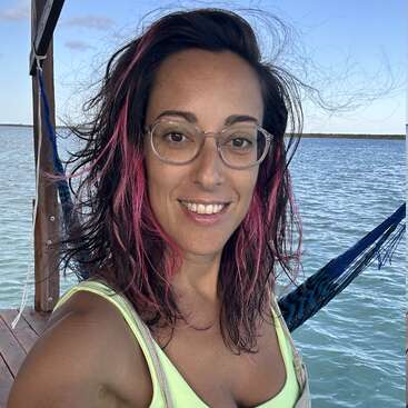 Una mujer con gafas y el pelo a la altura de los hombros con mechas rosas sonríe a la cámara. Está de pie junto al agua; se ven una hamaca y un embarcadero de madera.
