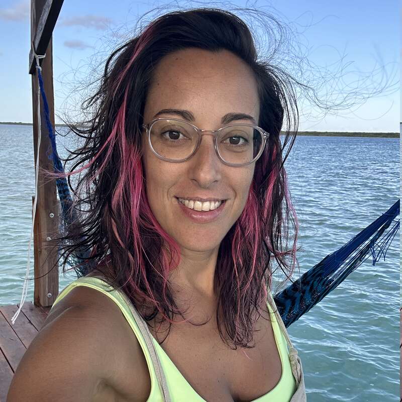 Una mujer con gafas y el pelo a la altura de los hombros con mechas rosas sonríe a la cámara. Está de pie junto al agua; se ven una hamaca y un embarcadero de madera.