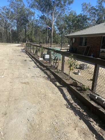 L'image représente une clôture rustique avec une maison et des arbres à l'arrière-plan, avec un chemin de terre et un mât arborant le drapeau australien.