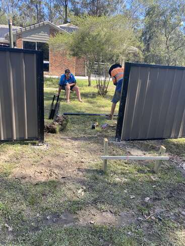 L'image représente deux hommes construisant une clôture dans une cour, l'un assis dans l'herbe et l'autre debout, entouré d'outils et de matériaux.