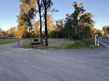 Une entrée rurale avec un portail métallique, des arbres et une route pavée. La lumière du soleil filtre à travers la forêt. Une maison est visible au loin. Atmosphère paisible.