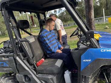Un jeune homme vêtu d'un sweat à capuche à rayures bleues dort dans un véhicule utilitaire 4x4, tandis qu'un autre homme se tient à l'extérieur. Autour d'eux, des arbres et de l'herbe.