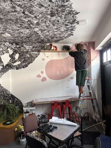 A person stands on a ladder, arranging objects on a shelf in a creatively decorated room with collage art on the ceiling and eclectic furniture scattered below.