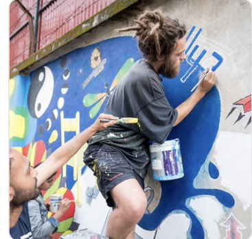 The image depicts a group of people painting a mural on a wall, showcasing their artistic skills and creativity. The mural features a rainbow and human figures.