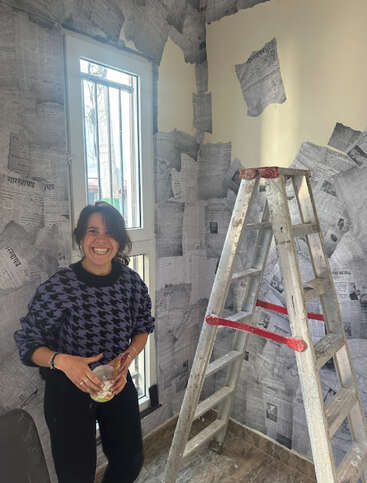 A smiling woman stands beside a ladder, holding a container. The walls are being decorated with newspaper clippings, creating a creative, unique wallpaper effect in the room.
