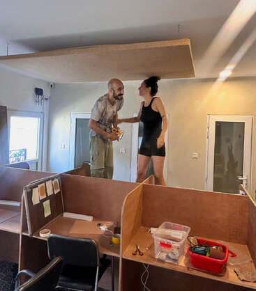 A man and woman stand laughing on a desk in an office, surrounded by tools and materials, appearing to be working on a ceiling panel project.