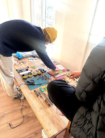 Two people are working together at a sunlit table filled with assorted electronic components, tools, wires, and gadgets, possibly assembling or repairing circuits in a cozy setting.
