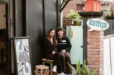 Two people sit closely together outside a cozy café with plants and artistic décor. A sign reads "Sattva" and another board displays "Art Café" artwork.