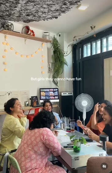 A group of women are laughing and enjoying themselves around a table in a cozy room, decorated with plants and fairy lights. Text reads: “But girls they wanna have fun.”