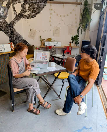 Two people sit at a small café table, engaged in conversation. The setting is cozy, decorated with plants, string lights, and creative wall art, creating a warm atmosphere.