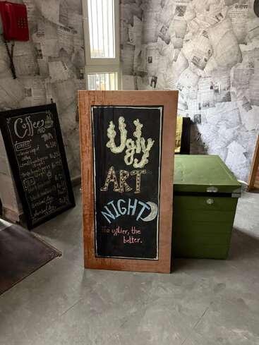 This image shows a quirky cafe with a chalkboard sign advertising “Ugly Art Night: The uglier, the better.” A coffee menu and newspaper-covered walls are visible.
