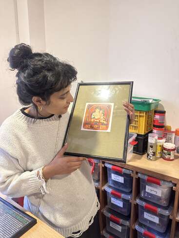 A woman stands indoors, holding and admiring a framed artwork featuring a deity. Behind her are organized bins and various supplies labeled in a creative workspace.