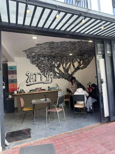 A cozy, open-front café named "Sattya" features a striking tree mural; people sit and work inside, surrounded by eclectic furniture and creative, relaxed vibes.