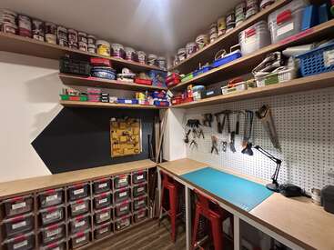 This image shows a well-organized workshop with shelves full of supplies, labeled storage bins, a pegboard with tools, a workbench, stools, and ample lighting.