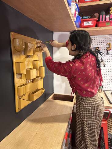 A woman in a red patterned top installs a yellow wall organizer on a black wall in a workshop, surrounded by shelves and storage boxes.