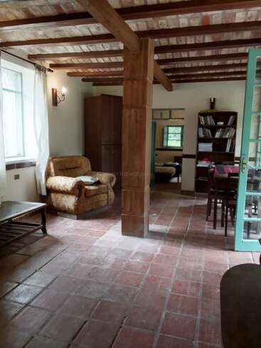 This cozy room features rustic brick floors and wooden beams, an armchair near a window, bookshelf, wardrobe, coffee table, dining area, and natural light.
