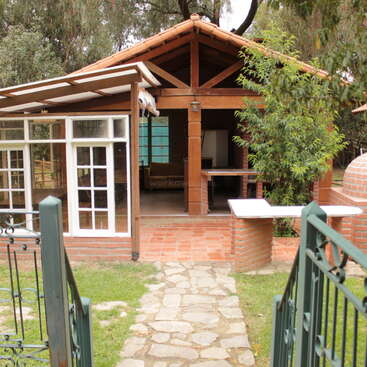 A small rustic house with a red tiled roof, wooden beams, brick walls, and a glass sunroom, surrounded by greenery, viewed from an open gate.