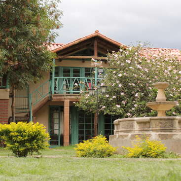 Ein hübsches Haus mit Ziegeldach, grünem Holzbalkon, blühenden rosa Blumen, Springbrunnen und üppigem Garten, das unter einem bewölkten Himmel eine ruhige, einladende Atmosphäre schafft.