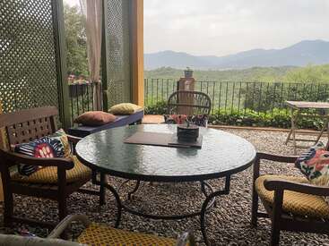 Eine gemütliche Terrasse mit einem runden Glastisch, Holzstühlen, bunten Kissen, Kieselsteinboden, Gitterwänden, Bergblick, viel Grün und sanftem Licht schafft einen Ort der Ruhe.