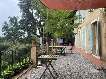 Eine gemütliche Terrasse im Freien mit Holzmöbeln, Kiesboden, üppigem Grün, Bäumen, Topfpflanzen und einem charmanten gelben Haus mit blauen Fensterläden und einem Überdachung.