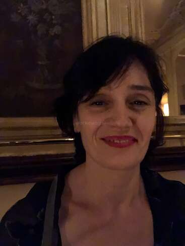 Eine Frau mit kurzen dunklen Haaren lächelt die Kamera herzlich an. Die Kulisse ist schwach beleuchtet, elegant eingerichtet und im Hintergrund hängt ein Gemälde.