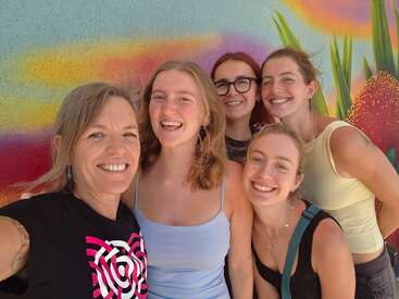 Fünf lächelnde Frauen machen zusammen ein Selfie vor einem bunten, lebendigen Wandbild mit Pflanzenmotiven. Sie sehen glücklich, fröhlich und wie enge Freundinnen aus.