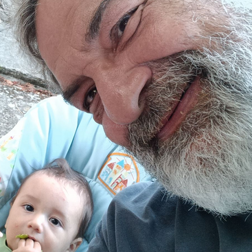 Um homem barbudo sorri para a câmera, enquanto um bebê está sentado em uma cadeirinha azul, olhando para cima e segurando comida. Os dois parecem felizes e relaxados ao ar livre.