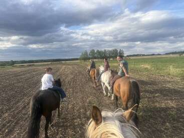 A group of people, including children, ride horses through an open field under a cloudy sky. The scene is peaceful, rural, and surrounded by nature.
