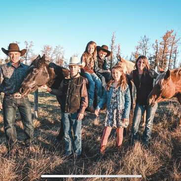A family of six stands outdoors in a field with two horses. They wear western clothes and cowboy hats, smiling under a clear blue sky.