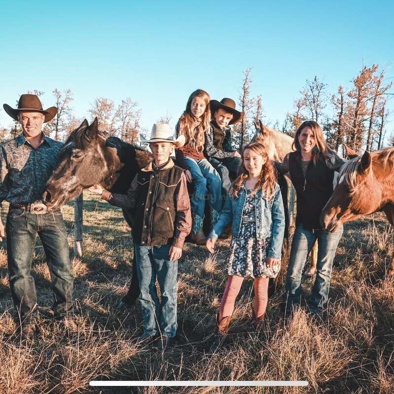 A family of six stands outdoors in a field with two horses. They wear western clothes and cowboy hats, smiling under a clear blue sky.