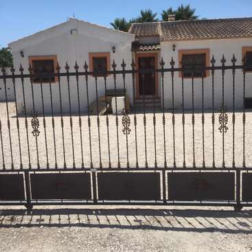 La imagen muestra una casa blanca con tejado de tejas, con una valla metálica negra en primer plano y un cielo azul despejado.