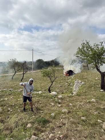 Una persona vestida con una sudadera con capucha y pantalones cortos excava en un terreno rocoso con una pala, mientras que, al fondo, una pila de escombros en llamas desprende un humo espeso.