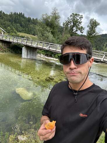 A man wearing large sunglasses stands by a clear river, holding food. Behind him is a wooden bridge, green trees, and cloudy skies in the background.