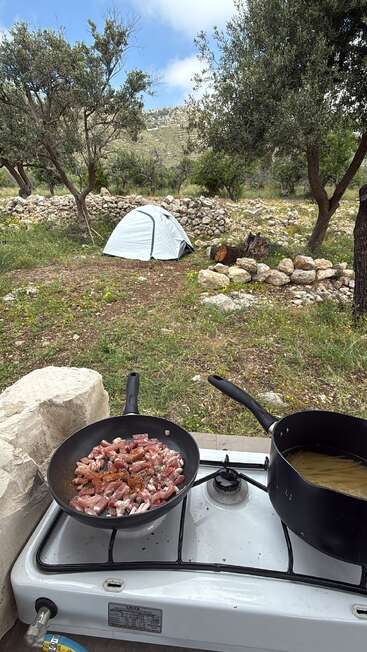 A small tent stands among trees and rocks. In the foreground, a camping stove cooks pasta and diced meat, creating a peaceful outdoor cooking scene.