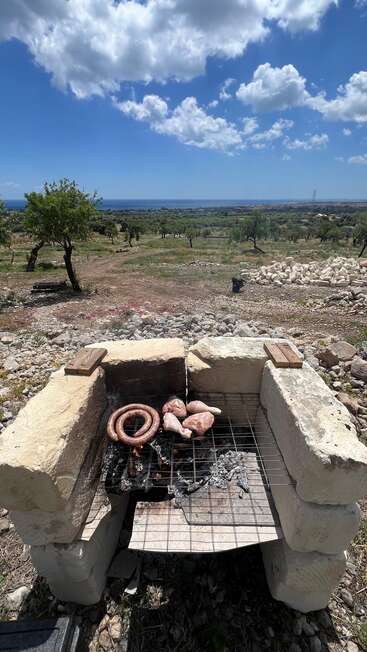 A rustic outdoor barbecue grill made of stone stands in a rocky field, cooking sausage and meat under a bright blue sky with fluffy clouds. Peaceful countryside.