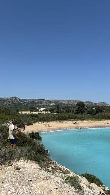 A man stands on rocky terrain overlooking a beautiful, turquoise bay and sandy beach with a few people. Beyond, lush greenery and distant mountains under clear sky.