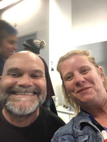 Un homme et une femme sourient pour un selfie pris à l'intérieur. On aperçoit deux personnes à l'arrière-plan. L'ambiance est décontractée et conviviale.
