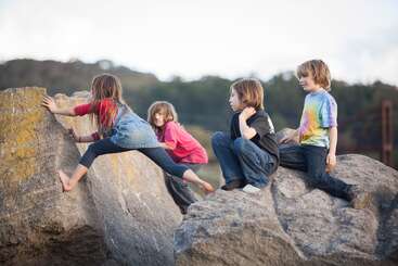 Quatre enfants sont dehors ; ils grimpent et sont assis sur de gros rochers. Une petite fille grimpe, tandis que les trois autres la regardent. À l'arrière-plan, on voit des arbres et le ciel.