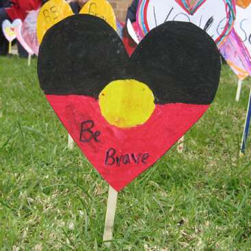 Un panneau en forme de cœur, peint aux couleurs du drapeau aborigène et portant l'inscription « Be Brave », posé dans l'herbe, avec d'autres panneaux en forme de cœur colorés en arrière-plan.