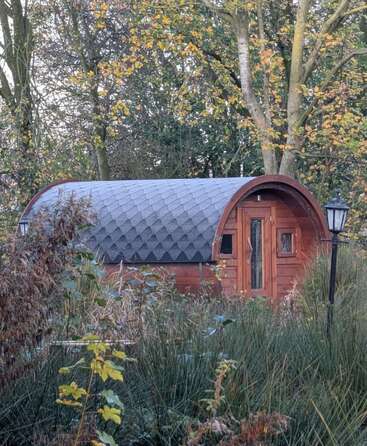 Eine kleine, tonnenförmige Holzhütte mit Schindeldach steht inmitten von hohen Gräsern und herbstlichen Bäumen, neben einem klassischen schwarzen Laternenpfahl.