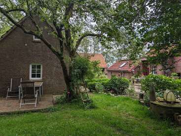 Ein gemütlicher Hinterhof mit grünem Gras, Bäumen, einem Holztisch und Stühlen, einer Leiter an einem Baum und charmanten Backsteinhäusern mit roten Ziegeldächern.