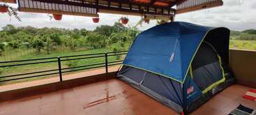 Ein blaues Zelt steht auf einer überdachten Terrasse mit Blick auf eine üppige grüne Landschaft. Hängende Töpfe und Grünpflanzen schaffen eine friedliche Camping-Atmosphäre im Freien.