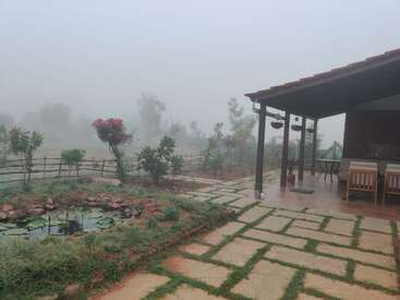Ein nebliger Morgen in einem Haus auf dem Land mit einer gefliesten Veranda, Stühlen im Freien, einem Seerosenteich, Pflastersteinen, blühenden Pflanzen und fernen Bäumen, die im Nebel kaum zu erkennen sind.