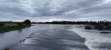 Ein ruhiger Fluss fließt über einen niedrigen Kaskadendamm, gesäumt von üppigem Grün unter einem wolkenverhangenen Himmel, mit fernen Bäumen, die sich im schwindenden Abendlicht abzeichnen.