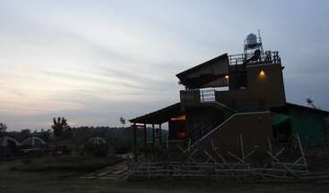 A two-story building with an observatory dome on the roof, surrounded by tents and greenery at dusk. The sky is cloudy, creating a tranquil atmosphere.