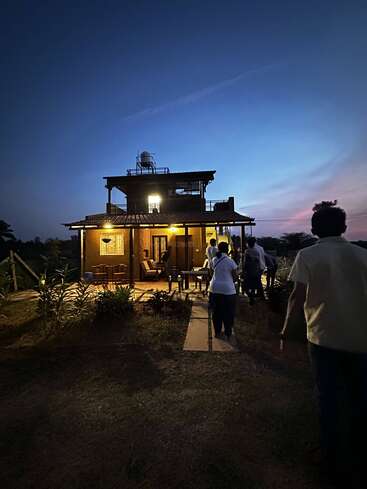 A group of people walk towards a warmly lit, rustic house at dusk. The sky is deep blue, creating a peaceful, inviting evening atmosphere around them.