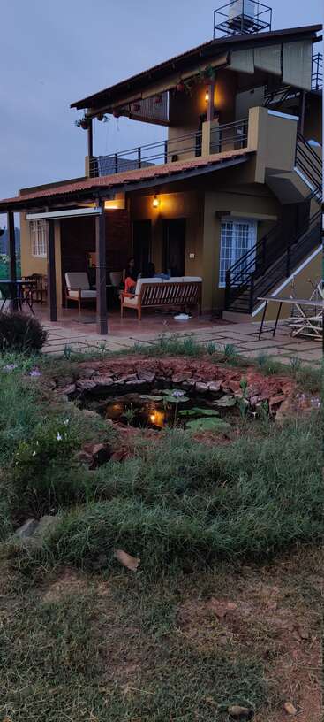 A cozy two-story house with a patio, warm lights glowing, surrounded by nature. There’s a small pond with water lilies and lush green grass.