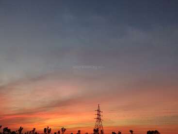 A beautiful sunset sky with vibrant orange, pink, and blue hues. Silhouettes of trees and an electrical tower create a peaceful, serene landscape scene.