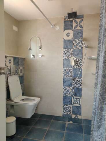 This image shows a modern bathroom featuring a wall-mounted toilet, overhead shower, handheld shower, blue patterned tiles, glass shelves, blue floor tiles, and a small trash bin.